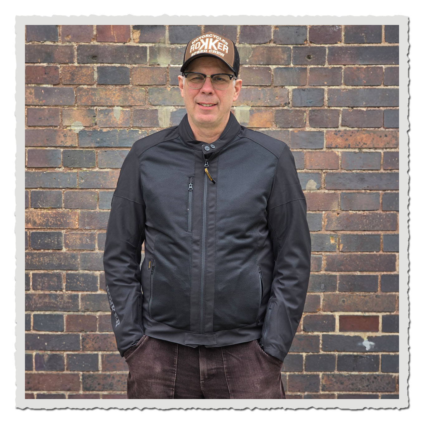 Mann in schwarzer Jacke und brauner Kappe, Frontalansicht vor einer braunen Backsteinwand.